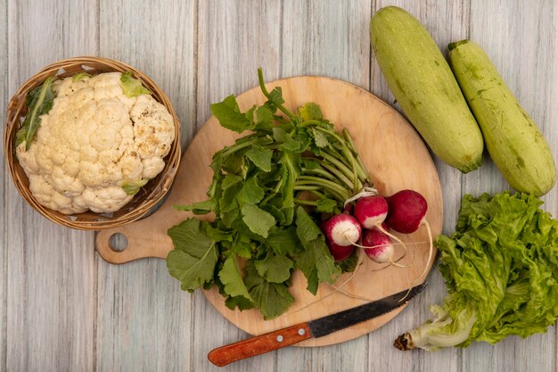 Vista dall'alto di ravanelli freschi su una tavola da cucina in legno con coltello con cavolfiore su un secchio con lattuga e zucchine isolato su una parete in legno grigio