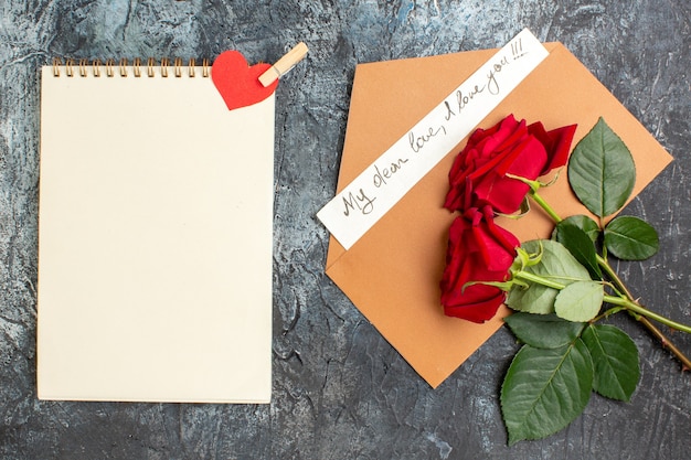 Vista dall'alto di rose rosse e busta con lettera d'amore e quaderno a spirale su sfondo scuro ghiacciato