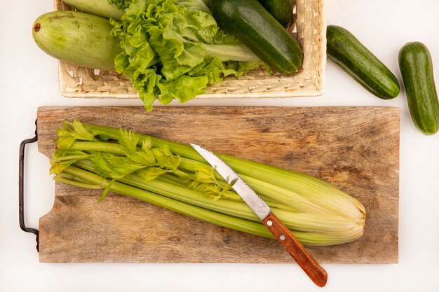 Vista dall'alto di sedano fresco su una tavola da cucina in legno con coltello con cetrioli zucchine e lattuga su un secchio su una superficie bianca