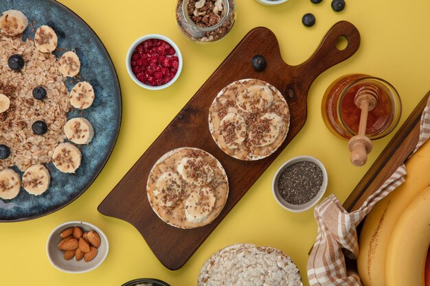 Vista dall'alto di snack con banana burro di arachidi sesamo su pane croccante sul tagliere con marmellata di mandorle di ribes rosso prugnolo e farina d'avena su sfondo giallo