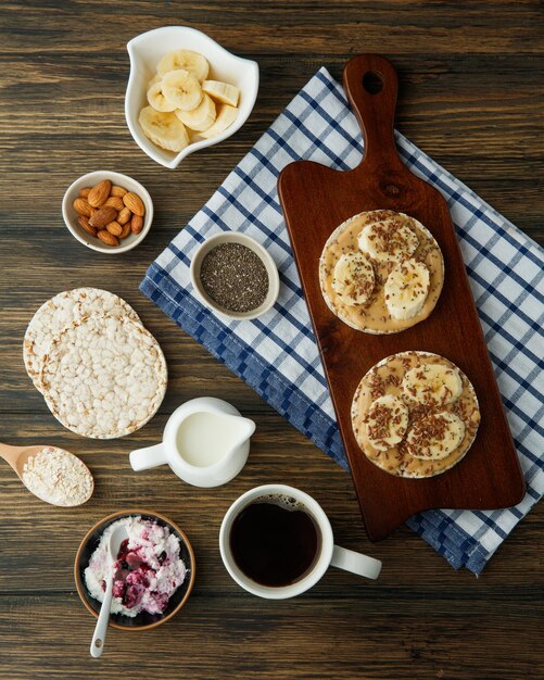 Vista dall'alto di snack di pane croccante al burro di arachidi e banana sul tagliere con pane croccante al latte di mandorle e caffè di avena su panno a quadri su fondo di legno