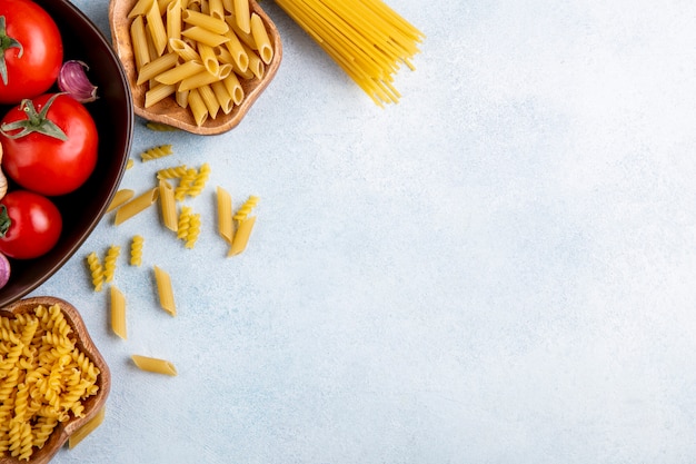 Vista dall'alto di spaghetti crudi con pasta cruda in ciotole con aglio e pomodori in una ciotola su una superficie grigia