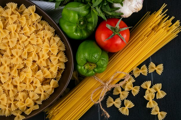 Vista dall'alto di spaghetti crudi con pasta cruda in una ciotola con pomodori, aglio e peperone su una superficie nera