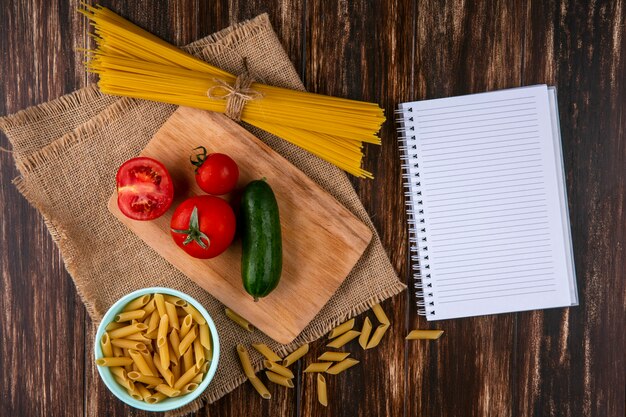 Vista dall'alto di spaghetti crudi con pomodori e cetrioli su un tagliere con un taccuino su un tovagliolo beige su una superficie di legno