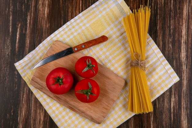 Vista dall'alto di spaghetti crudi con pomodori su un tagliere con un coltello su un asciugamano a scacchi giallo su una superficie di legno