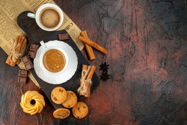 Vista dall'alto di tazze di caffè su tagliere di legno e un vecchio giornale biscotti cannella lime barrette di cioccolato sul lato destro su sfondo scuro