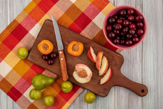 Vista dall'alto di tutta la frutta tagliata e affettata come ciliegia albicocca pesca con coltello sul tagliere e ciliegie in una ciotola con pera e prugne su panno plaid su sfondo di legno