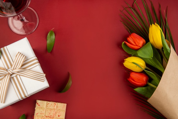 Vista dall'alto di un bouquet di tulipani di colore rosso e giallo con un bicchiere di vino, cioccolata bianca e una confezione regalo sul tavolo rosso con spazio di copia