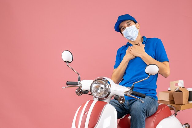 Vista dall'alto di un corriere grato in maschera medica che indossa un cappello seduto su uno scooter su una pesca pastello