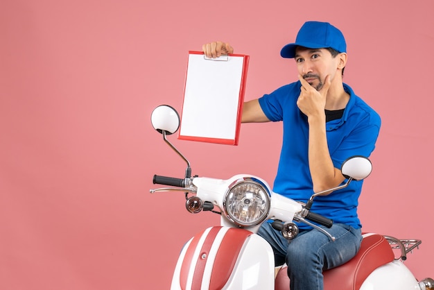 Vista dall'alto di un corriere pensante che indossa un cappello seduto su uno scooter che tiene un documento su una pesca pastello