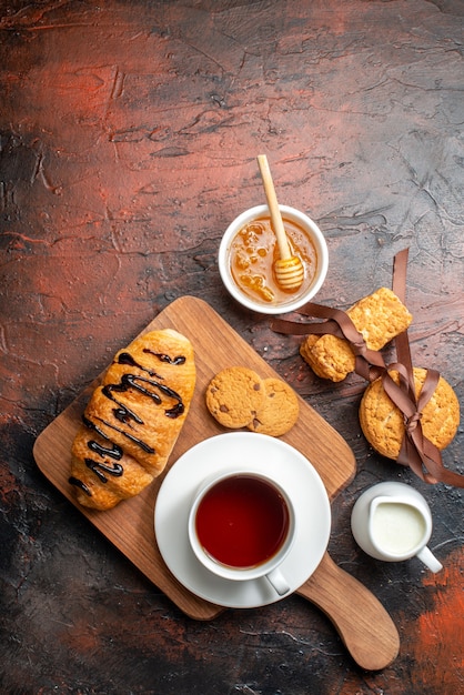 Vista dall'alto di un delizioso croissant una tazza di tè nero su un tagliere di legno biscotti impilati miele latte su una superficie scura dark
