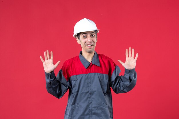 Vista dall'alto di un giovane costruttore sorridente in uniforme che indossa un elmetto che mostra dieci sul muro rosso isolato