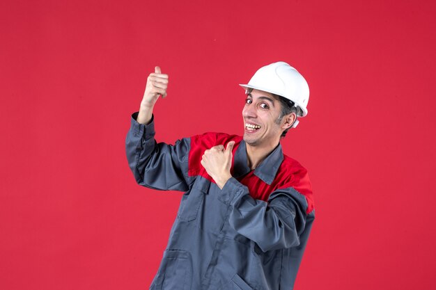 Vista dall'alto di un giovane costruttore sorridente in uniforme con elmetto e che fa un gesto ok sul muro rosso isolato