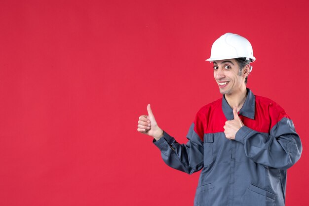 Vista dall'alto di un giovane lavoratore sorridente fiducioso in uniforme con elmetto e che fa un gesto ok sul muro rosso isolato