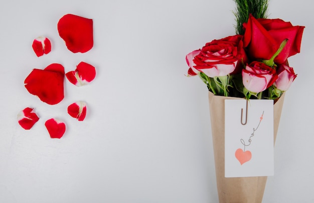 Vista dall'alto di un mazzo di rose di colore rosso con asparagi in carta artigianale con allegata cartolina con una graffetta e petali di fiori rossi su sfondo bianco con spazio di copia