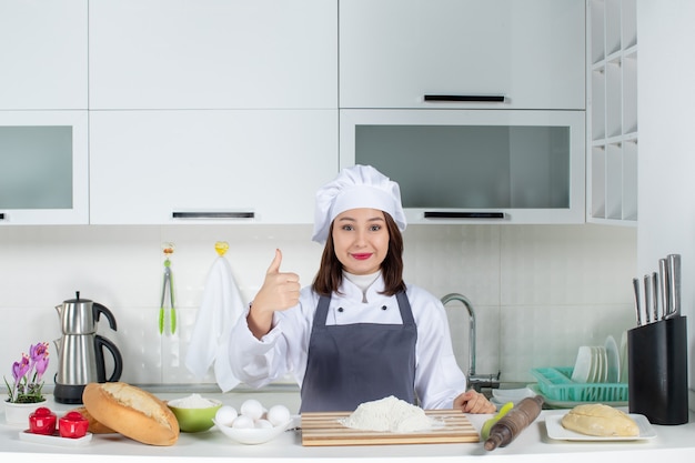 Vista dall'alto di una chef donna sicura di sé in uniforme in piedi dietro il tavolo con tagliere di pane e verdure che fa un gesto ok nella cucina bianca