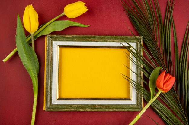 Vista dall'alto di una cornice vuota e tulipani di colore giallo e rosso con foglia di palma sul tavolo rosso