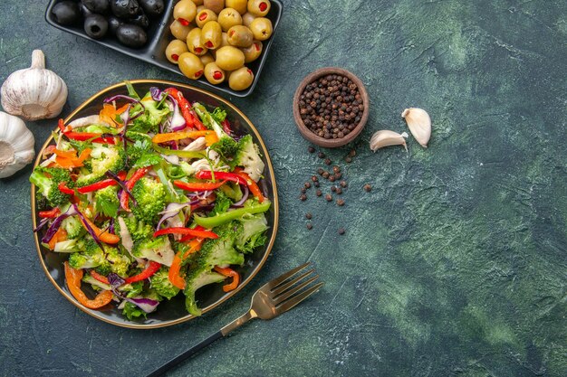 Vista dall'alto di una deliziosa insalata vegana in un piatto con varie verdure e forchetta pepe verde olive nere aglio sul lato destro su sfondo scuro