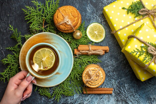 Vista dall'alto di una deliziosa torta cremosa e rami di abete limone cannella lime scatole regalo gialle mano che tiene tazza di tè nero su sfondo scuro