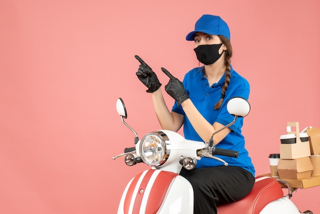 Vista dall'alto di una donna corriere sicura che indossa maschera medica e guanti seduti su uno scooter che consegna ordini su sfondo color pesca pastello