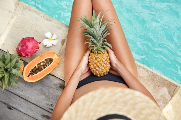 Vista dall'alto di una donna magra con la pelle abbronzata, si siede vicino alla piscina dell'hotel, gode di una dieta sana vegana e mangia frutta tropicale, ha una festa in piscina estiva. La femmina mangia succosa frutta esotica: ananas e papia