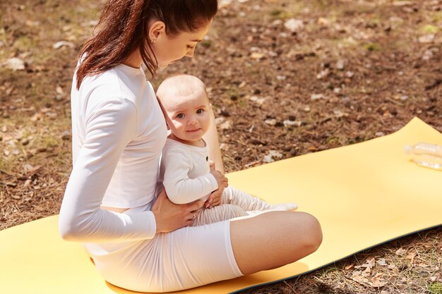 Vista dall'alto di una donna sportiva con un bambino seduto su un karemat nella posa del loto, tenendo le gambe incrociate