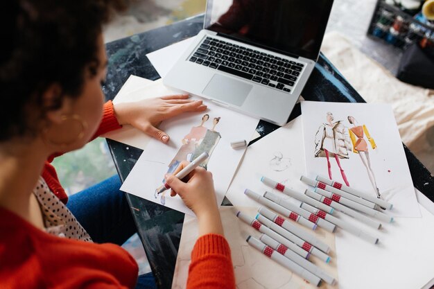 Vista dall'alto di una giovane donna con i capelli ricci scuri seduta al tavolo con un laptop che disegna sognante illustrazioni di moda in un'officina moderna