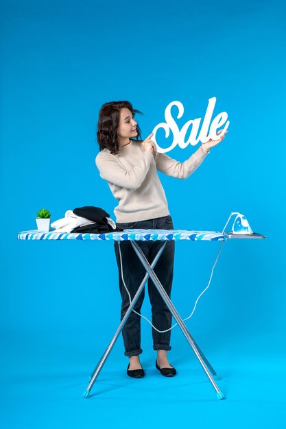 Vista dall'alto di una giovane donna sicura di sé in piedi dietro l'asse da stiro e che mostra l'icona di vendita su sfondo blu