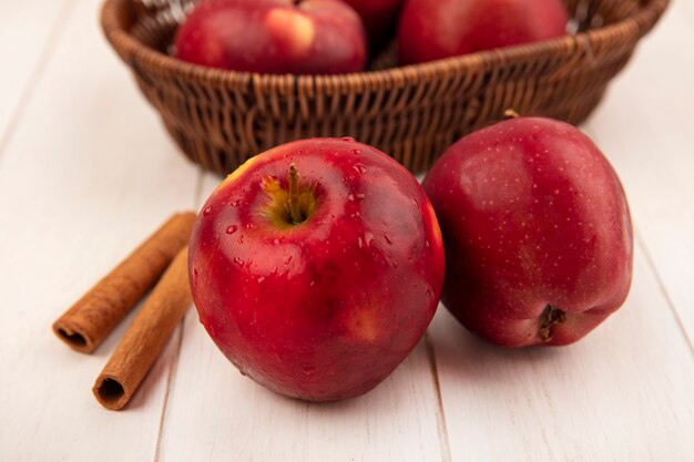 Vista dall'alto di una mela rossa con le mele su un secchio con bastoncini di cannella isolato su una superficie di legno bianca