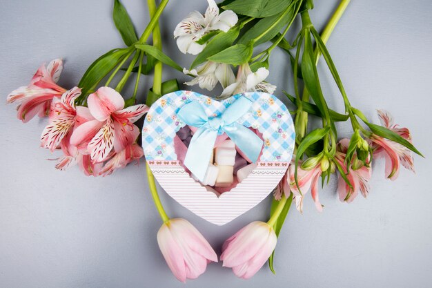 Vista dall'alto di una scatola presente a forma di cuore con marshmallow e alstroemeria di colore rosa e bianco e fiori di tulipano sul tavolo bianco