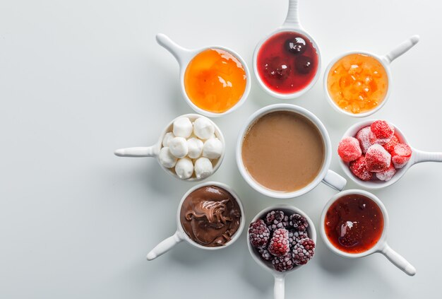 Vista dall'alto di una tazza di caffè con marmellate, lamponi, zucchero, cioccolato in tazze su superficie bianca