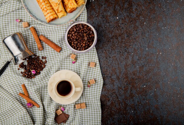 Vista dall'alto di una tazza di caffè e chicchi di caffè in una ciotola con bastoncini di cannella sulla tovaglia plaid con spazio di copia