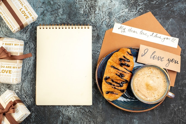 Vista dall'alto di una tazza di caffè e di un delizioso croissant fresco, bellissime scatole regalo per l'amato e quaderno a spirale su superficie scura