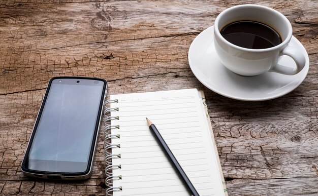 Vista dall&#39;alto di una tazza di caffè, telefono astuto e matita nera sul taccuino vuoto sopra il tavolo di legno.