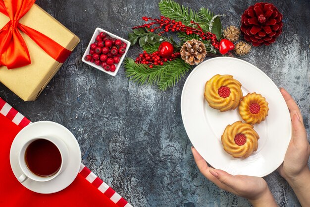 Vista dall'alto di una tazza di tè nero su un asciugamano rosso e biscotti in mano su un piatto bianco regalo di accessori di capodanno con nastro rosso su superficie scura