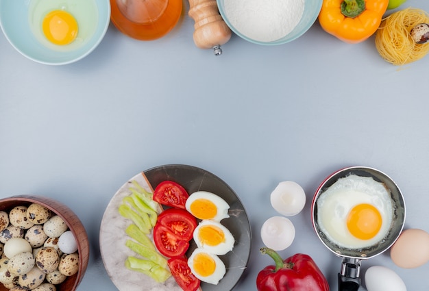 Vista dall'alto di uova di quaglia su una ciotola di legno con uova sode a metà su un piatto con fette di pomodoro con uova fritte su una padella su sfondo bianco con spazio di copia