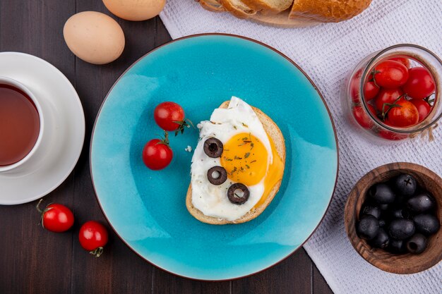 Vista dall'alto di uovo fritto con pomodori e olive nel piatto e ciotole di pomodoro e pane alle olive su stoffa con uova e tè su legno