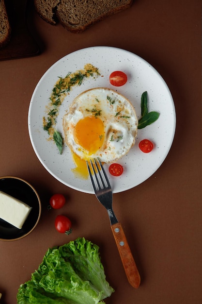 Vista dall'alto di uovo fritto con pomodoro e aneto con forchetta nel piatto e lattuga pomodori con burro su sfondo marrone