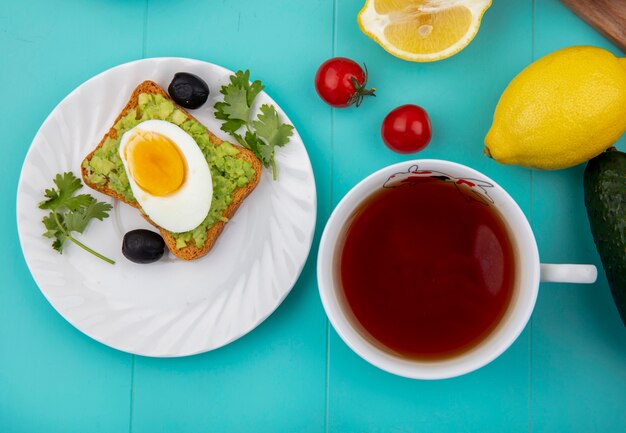 Vista dall'alto di uovo fritto su fetta di pane tostato con polpa di avocado sulla piastra bianca con olive nere con una tazza di tè sul blu