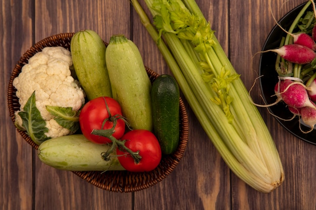 Vista dall'alto di verdure colorate come pomodori zucchine cetriolo e cavolfiore su un secchio con ravanelli su una ciotola con sedano isolato su una parete in legno