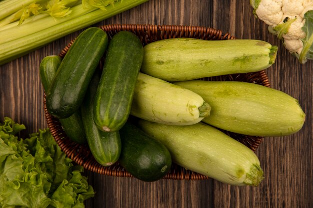 Vista dall'alto di verdure fresche a foglia verde come zucchine e cetrioli su un secchio con lattuga, sedano e cavolfiore isolato su una parete in legno