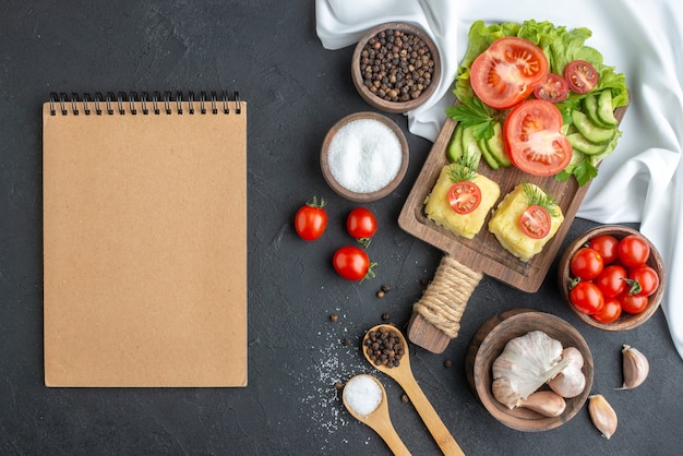 Vista dall'alto di verdure fresche tritate e intere sul tagliere in ciotole e spezie notebook su asciugamano bianco su superficie nera