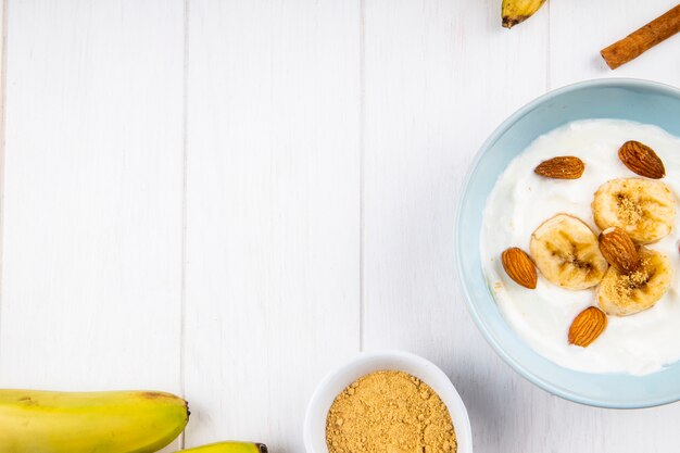 Vista dall'alto di yogurt con banana e mandorle, bastoncini di cannella su bianco con spazio di copia