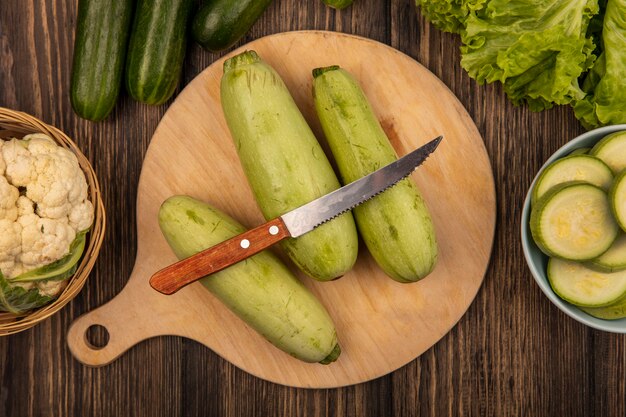 Vista dall'alto di zucchine isolato su una tavola da cucina in legno con coltello con cavolfiore su un secchio con cetrioli e lattuga isolato su una superficie in legno