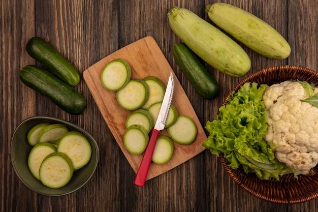 Vista dall'alto di zucchine tritate su una ciotola con zucchine tritate su una tavola da cucina in legno con coltello con cavolfiore e lattuga su un secchio con cetriolo e zucchine isolato su una parete in legno