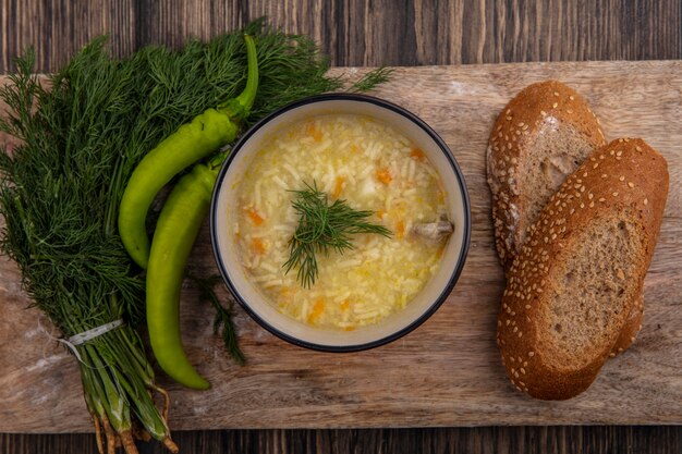 Vista dall'alto di zuppa di orzo di pollo nella ciotola e seminate di pannocchia marrone fette di pane con aneto e pepe sul tagliere su sfondo di legno