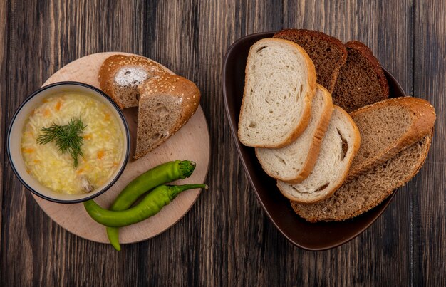 Vista dall'alto di zuppa di pollo orzo nella ciotola e peperoni sul tagliere con semi di pannocchia marrone bianco e fette di pane di segale nella ciotola su sfondo di legno