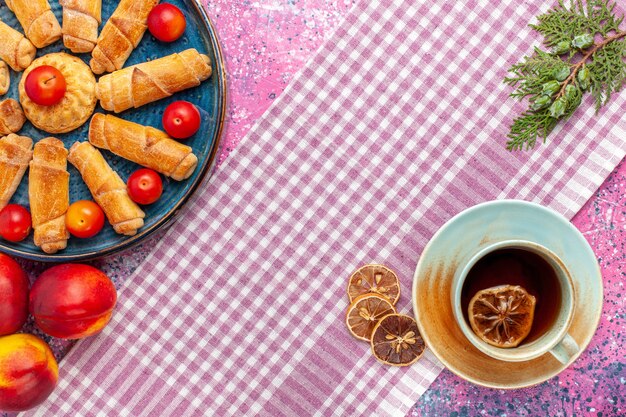 Vista dall'alto dolci deliziosi bagel all'interno del vassoio con prugne pesche e tazza di tè sulla scrivania rosa chiaro