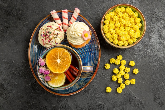 Vista dall'alto dolci sul piatto cupcakes con crema bianca sul piattino una tazza di tè con limone e bastoncini di cannella ciotola di dolci gialli sul tavolo