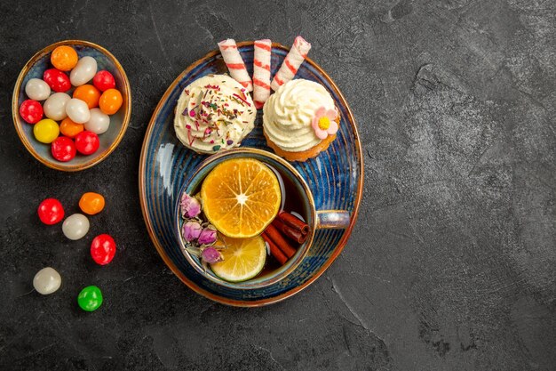 Vista dall'alto dolci sul piatto due appetitosi cupcakes e dolci sul piattino una tazza di tè con limone e bastoncini di cannella ciotole di dolci colorati sul tavolo